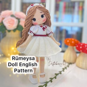 Puede incluir: Muñeca de ganchillo hecha a mano con pelo largo castaño, vestida con un vestido blanco con detalles rojos, una diadema a juego y zapatos blancos. La muñeca tiene una expresión dulce. El texto "Rümeysa Doll English Pattern" es visible.