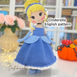 Może przedstawiać: Szydełkowa lalka Kopciuszka w niebieskiej sukience z białym obszyciem. Lalka ma blond włosy, niebieską opaskę i niebieskie rękawiczki. Widoczny tekst "Cinderella English pattern".