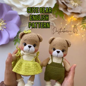 以下が含まれることがあります： 緑色のドレスを着たクマとオーバーオールを着たクマの2体の編みぐるみクマ。どちらも茶色の毛並み、白い顔、ピンクの頬をしています。クマたちは手をつないで、白い表面に座っています。クマたちの頭上に「Cute Bear English Pattern」という文字が書かれています。
