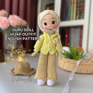 May include: A crocheted doll wearing a yellow shirt, brown pants, and a white headscarf. The doll is standing on a white surface with a wicker basket and a white lace ribbon in the background. The text "DURU DOLL HIJAP OUTFIT ENGLISH PATTERN" is visible in the image.