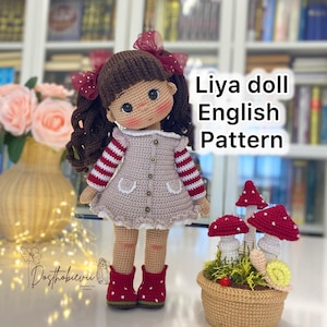 May include: A handmade crochet doll with brown hair and red bows, wearing a pink dress and red boots. The doll is next to a crochet basket with red mushroom decorations. Text on the image reads "Liya doll English Pattern".