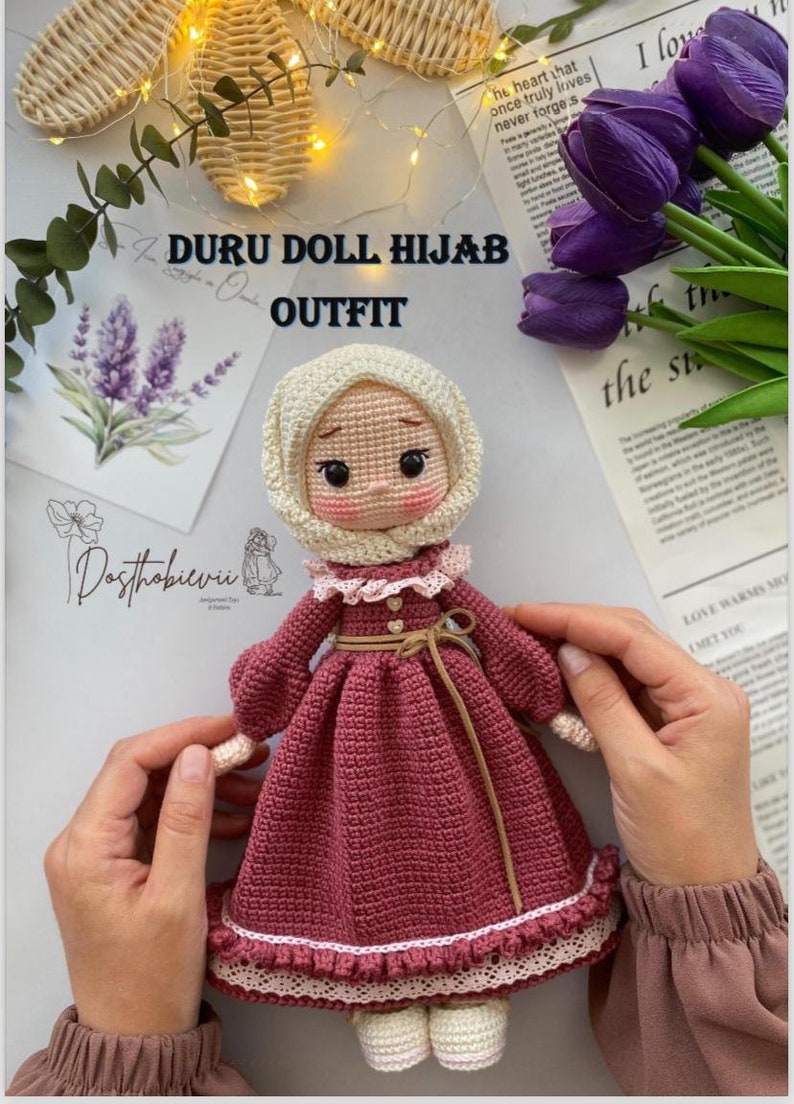 May include: A crocheted doll wearing a pink dress with a white lace trim, a white crocheted hijab, and brown shoes. The doll is holding a small crocheted flower in its hand. The doll is sitting on a white surface with a floral pattern.