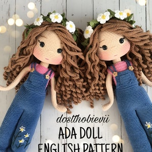 Könnte beinhalten: Zwei gehäkelte Puppen mit braunen Locken, die blaue Latzhosen und Blumenkränze tragen. Die Puppen liegen auf einer weißen Holzoberfläche. Der Text "dostthobievii ADA DOLL ENGLISH PATTERN" steht unter den Puppen.