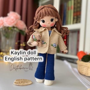 May include: Crochet doll pattern for a girl doll with brown hair, a beige jacket with floral embroidery, blue jeans, and white shoes. The doll is holding a small crocheted teddy bear. The pattern is called "Kaylin doll English pattern".