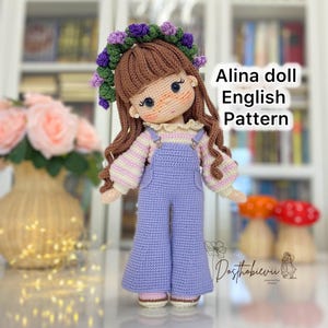 Può includere: Una bambola all'uncinetto fatta a mano con capelli castani, che indossa una tuta viola e una corona floreale. La bambola ha un incarnato chiaro e indossa una camicia a righe. Il testo "Alina doll English Pattern" è visibile. La bambola è in piedi davanti a uno sfondo sfocato.
