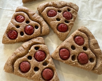 May include: Five homemade pizza-shaped dog treats with red pepperoni-shaped decorations on a white parchment paper background.