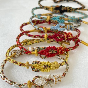 Könnte beinhalten: Eine Sammlung von sieben geflochtenen Armbändern, die in einer Reihe angeordnet sind und jeweils einen zentralen Zierknoten aufweisen. Die Armbänder bestehen aus mehrfarbigen Fäden in Gold, Rot, Weiß, Schwarz und Blau mit goldenen Akzenten.