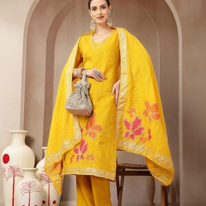 May include: A bright yellow salwar kameez set. The ensemble includes a tunic, pants, and a matching dupatta. The tunic and dupatta feature floral embroidery in shades of orange and pink. The outfit is accessorized with a silver beaded bag and earrings.