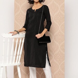 May include: A black tunic with embroidered detailing, worn with white trousers. The tunic features a split neckline and three-quarter sleeves. A black handbag with a silver chain is carried. The outfit is finished with silver-toned flats.