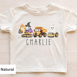 May include: A natural-colored short-sleeve t-shirt featuring a graphic of three yellow construction vehicles dressed for Halloween: an excavator with a witch hat, a dump truck, and a steamroller covered by a ghost sheet, with the name CHARLIE below.
