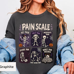 May include: A graphite-colored crewneck sweatshirt featuring a graphic print titled PAIN SCALE. The design displays six illustrated skeletons in various states of distress, labeled from NO PAIN to WORST PAIN POSSIBLE, with pumpkins, bats, and spiderwebs.