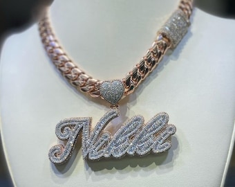 May include: A rose gold chain necklace with a large, custom name pendant. The pendant is set with clear cubic zirconia stones and features a heart-shaped charm at the top. The chain is a thick, curb style.