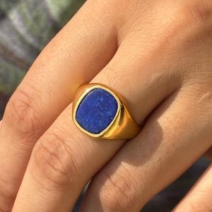 Puede incluir: Un anillo de sello de oro con una piedra preciosa azul. El anillo está en el dedo de una persona.