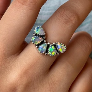 May include: A silver ring featuring a cluster of pear-shaped opals. The opals display a vibrant play-of-color with hues of green, blue, and yellow. The ring's design is a curved arrangement of the opals.