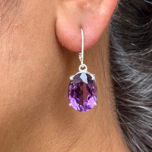 Könnte beinhalten: Nahaufnahme eines silbernen Ohrrings mit einem großen, oval geformten, tiefvioletten Amethyst-Edelstein. Der Edelstein ist in einer silbernen Lünette gefasst und hängt an einem silbernen Haken. Der Ohrring wird am Ohr einer Person getragen.