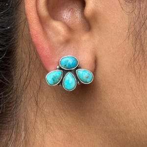 以下が含まれることがあります： ターコイズスタッドイヤリングのクローズアップ。イヤリングは、シルバーメタルにセットされた5つのターコイズストーンが特徴です。石は鮮やかな青緑色で、花のパターンに配置されています。イヤリングは耳に装着されています。
