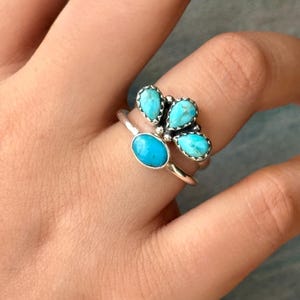 Könnte beinhalten: Zwei Silberringe mit Türkissteinen. Ein Ring hat einen ovalen Türkisstein, der andere vier tropfenförmige Türkissteine, die zusammengefasst sind. Die Ringe werden auf einer Hand präsentiert.