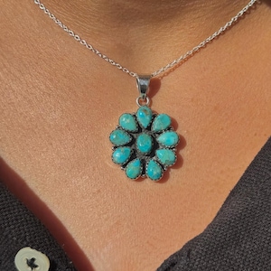 May include: A silver necklace with a pendant of turquoise stones in a floral design. The pendant hangs from a fine silver chain. The turquoise stones are a vivid blue-green. The necklace is worn against a dark background.