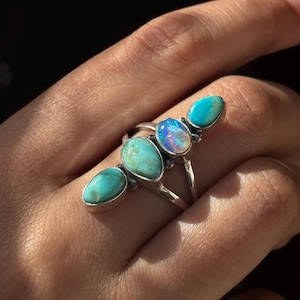 Op de afbeelding: Een zilveren ring met vijf turquoise en opaal stenen die in een aflopend patroon zijn gezet. De ring wordt gedragen aan een vinger.