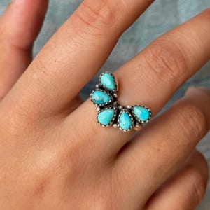 Può includere: Un anello d'argento con un design di pietre turchesi. L'anello ha una forma curva con più pietre turchesi incastonate in una fascia d'argento.