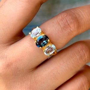 May include: A gold ring featuring three oval-cut gemstones. The ring has a central dark blue gemstone flanked by two lighter blue gemstones. The ring is set in a gold band, creating a striking contrast with the blue stones.