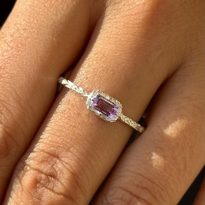 Puede incluir: Un anillo de plata con una banda delicada y texturizada y una pequeña piedra preciosa rectangular de color púrpura engastada en un engaste de plata.
