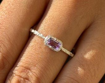Puede incluir: Un anillo de plata con una banda delicada y texturizada y una pequeña piedra preciosa rectangular de color púrpura engastada en un engaste de plata.