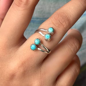 May include: A silver ring with three turquoise-colored stones. The ring has a unique design with the stones set in a scalloped bezel. The ring is worn on a finger, showcasing the jewelry's design and color.