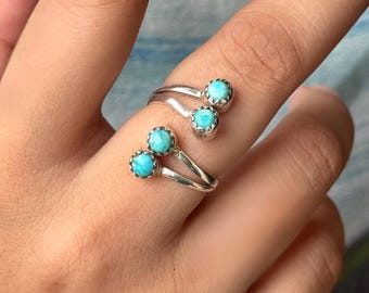 May include: A silver ring with three turquoise-colored stones. The ring has a unique design with the stones set in a scalloped bezel. The ring is worn on a finger, showcasing the jewelry's design and color.