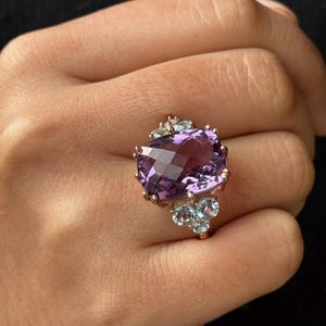 Op de afbeelding: Een ring van roségoud met een grote, gefacetteerde amethist-edelsteen, omringd door kleinere, gefacetteerde blauwe topazen.