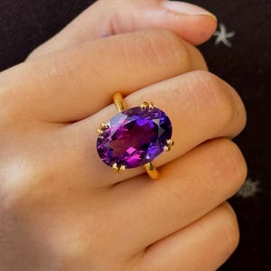 Könnte beinhalten: Ein goldener Ring mit einem großen, ovalen Amethyst-Edelstein. Der Edelstein ist tiefviolett und hat einen leichten Schimmer.