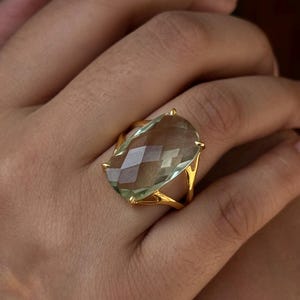 May include: A gold ring featuring a large, faceted, light green gemstone. The ring has a delicate, openwork design.