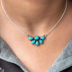 May include: A silver chain necklace featuring a central pendant composed of five teardrop-shaped turquoise stones set in silver bezels. The stones are arranged in a fan-like cluster, with one oval stone at the top center and four smaller stones below.