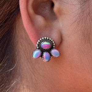 May include: A silver-toned stud earring featuring a cluster of iridescent oval-shaped stones. The central stone is surrounded by a beaded border, with four additional stones arranged below. The stones display a range of colors, including pink, blue, and green.