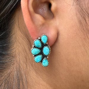 May include: A cluster stud earring featuring seven turquoise teardrop-shaped stones set in a silver-toned metal. The stones are a vibrant blue-green color, and the metal has a beaded edge.