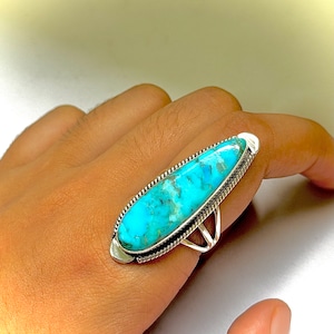 Peut inclure: Une bague en argent avec une grande pierre ovale en turquoise sertie dans une bélière en argent. La pierre est de couleur bleue et verte avec un motif marbré.