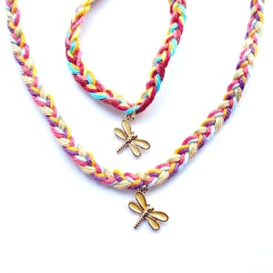 Könnte beinhalten: Ein geflochtenes Halsketten- und Armband-Set. Der Schmuck weist ein mehrfarbiges Muster aus rosa, gelben, blauen und cremefarbenen Fäden auf. Jedes Stück hat einen goldfarbenen Libellen-Anhänger. Das Set ist vor weißem Hintergrund.