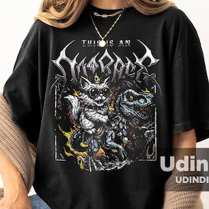 May include: Black graphic t-shirt featuring a detailed illustration of a cat-like creature and a dinosaur-like creature. The text "THIS IS AN" is above the illustration, with the word "SHIRRAGE" in a stylized font. The shirt is worn by a person.