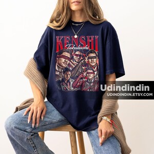 K&ouml;nnte beinhalten: Marineblaues T-Shirt mit einem Retro-Design. Der Text "Kenshi Takahashi" ist rot, und die Grafik zeigt Portr&auml;ts von Personen mit Augenbinden. Die Etsy-Shop-URL ist unten rechts sichtbar.