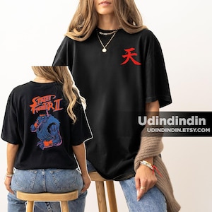 Puede incluir: Una camiseta negra de manga corta con un símbolo kanji rojo en el pecho. La parte posterior de la camiseta muestra un gráfico de un personaje en tonos azules de Street Fighter II con el texto naranja Street Fighter II encima.