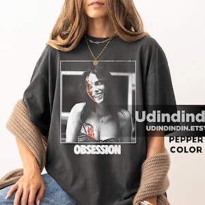 Könnte beinhalten: Ein anthrazitfarbenes Kurzarm-T-Shirt mit einem schwarz-weißen Grafikdruck einer lächelnden Person mit blutähnlichen Markierungen auf Gesicht und Brust. Das Wort OBSESSION ist in weißen Großbuchstaben unter dem Grafikbild aufgedruckt.
