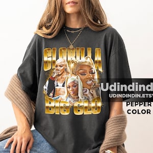 Puede incluir: Una camiseta de manga corta gris carbón con un estampado gráfico de la rapera GloRilla. El diseño incluye tres retratos fotográficos de la artista con el texto de color dorado GLORILLA y BIG GLO mostrado encima y debajo de las imágenes.