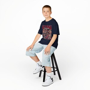 K&ouml;nnte beinhalten: Marineblaues T-Shirt mit rotem Schriftzug "KENSHI Takahashi" und Illustrationen von Figuren. Die Person tr&auml;gt hellblaue Jeansshorts und wei&szlig;e Turnschuhe und sitzt auf einem schwarzen Hocker.