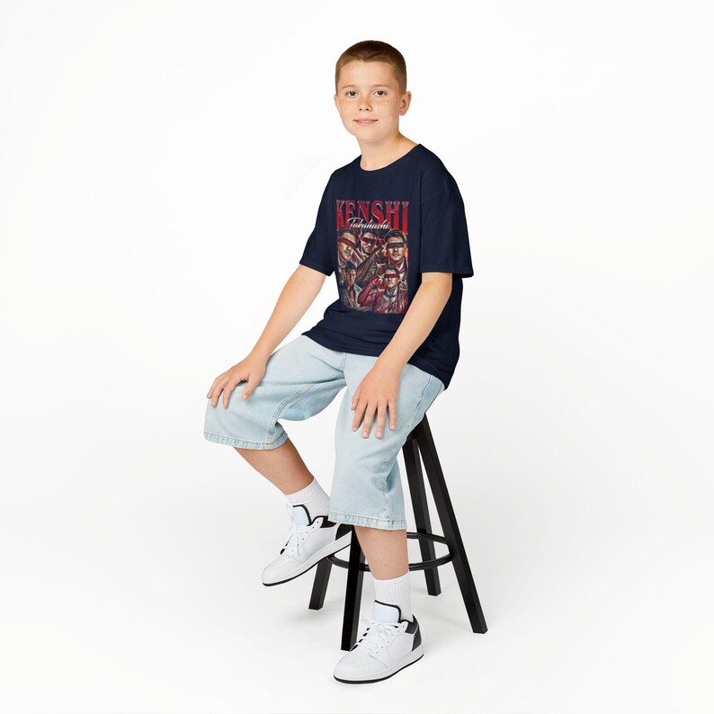 K&ouml;nnte beinhalten: Marineblaues T-Shirt mit rotem Schriftzug "KENSHI Takahashi" und Illustrationen von Figuren. Die Person tr&auml;gt hellblaue Jeansshorts und wei&szlig;e Turnschuhe und sitzt auf einem schwarzen Hocker.