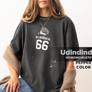 Może przedstawiać: Ciemnoszary t-shirt z krótkim rękawem z białym nadrukiem graficznym na klatce piersiowej. Nadruk przedstawia napis DI LAURENTIS nad liczbą 66 oraz stylizowaną ilustrację głowy i ramion osoby z profilu.