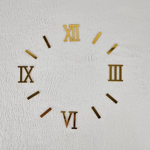 Può includere: Un muro bianco con numeri romani dorati di un orologio. I numeri sono XII, IX, III e VI.