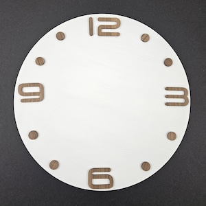 Op de afbeelding: Een witte wijzerplaat met houten cijfers op 12, 3, 6 en 9 uur. Kleine houten stippen markeren de andere uurposities. De wijzerplaat is rond en staat tegen een donkere achtergrond.