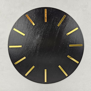 May include: A black, round clock face with gold-colored rectangular hour markers. The clock face has a textured surface and a minimalist design, suitable for modern decor.