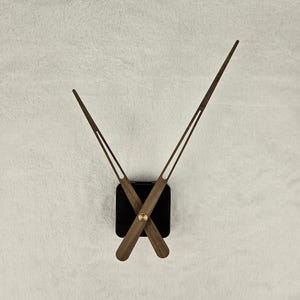 Op de afbeelding: Een klokmechanisme met houten wijzers en een zwarte vierkante basis. De uur- en minutenwijzers zijn bruin en slank, met een messingkleurig midden. De wijzers van de klok staan in een hoek, wat de tijd suggereert.