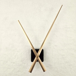 Op de afbeelding: Een moderne wandklok met een minimalistisch ontwerp. De klok heeft een zwarte vierkante basis en twee lichtbruine houten wijzers. De wijzers kruisen elkaar en de klok is bevestigd aan een witte, getextureerde muur.